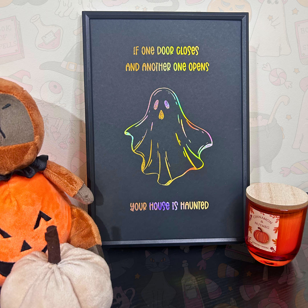 A framed Halloween art print with a black background, holographic ghost illustration, and the text 'IF ONE DOOR CLOSES AND ANOTHER ONE OPENS YOUR HOUSE IS HAUNTED'. The frame is styled on a dark tabletop next to a plush pumpkin and a candle.