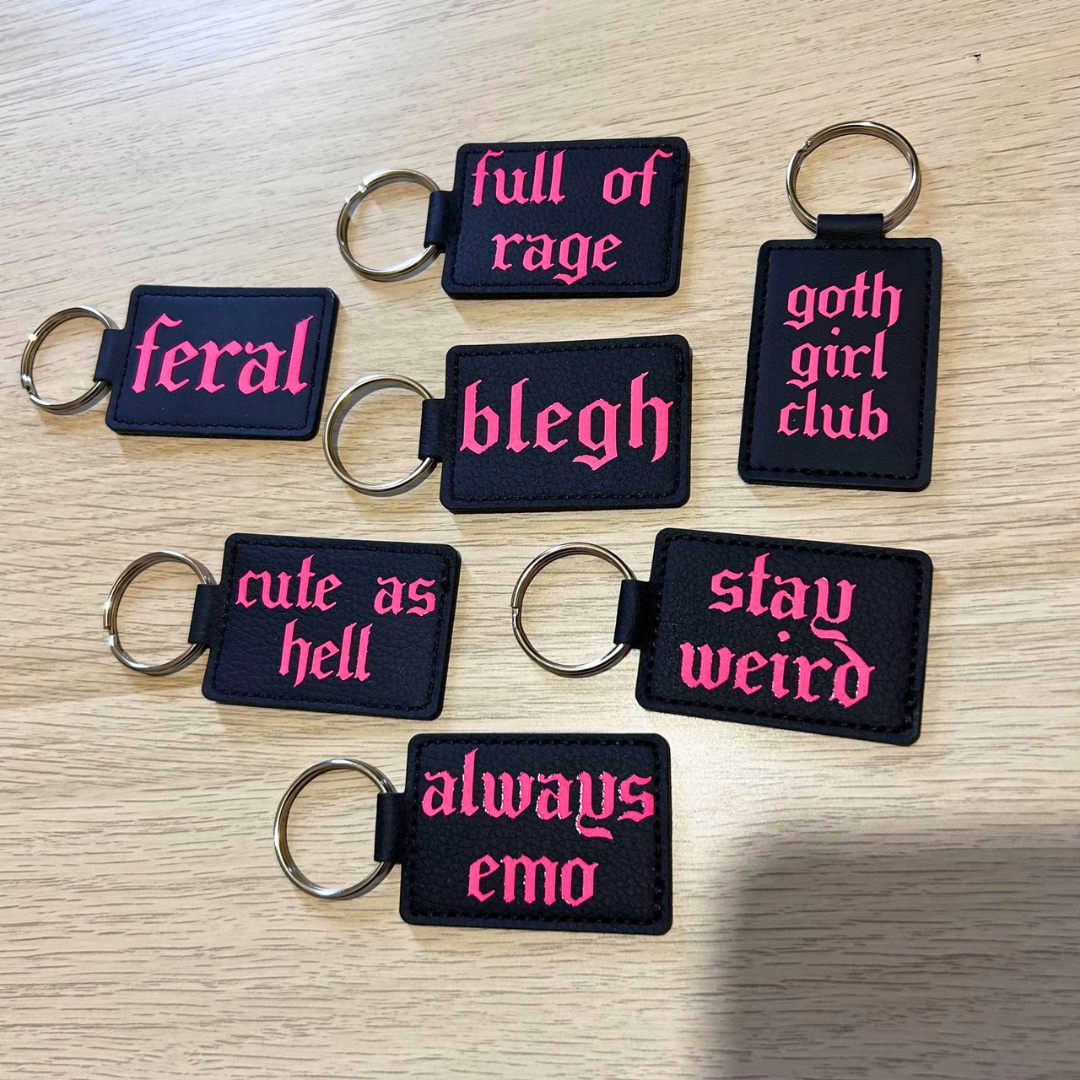 Seven black rectangular keychains with pink Gothic-style text are arranged on a light wood surface. The phrases include 'feral', 'full of rage', 'goth girl club', 'bleugh', 'cute as hell', 'stay weird', and 'always emo'.