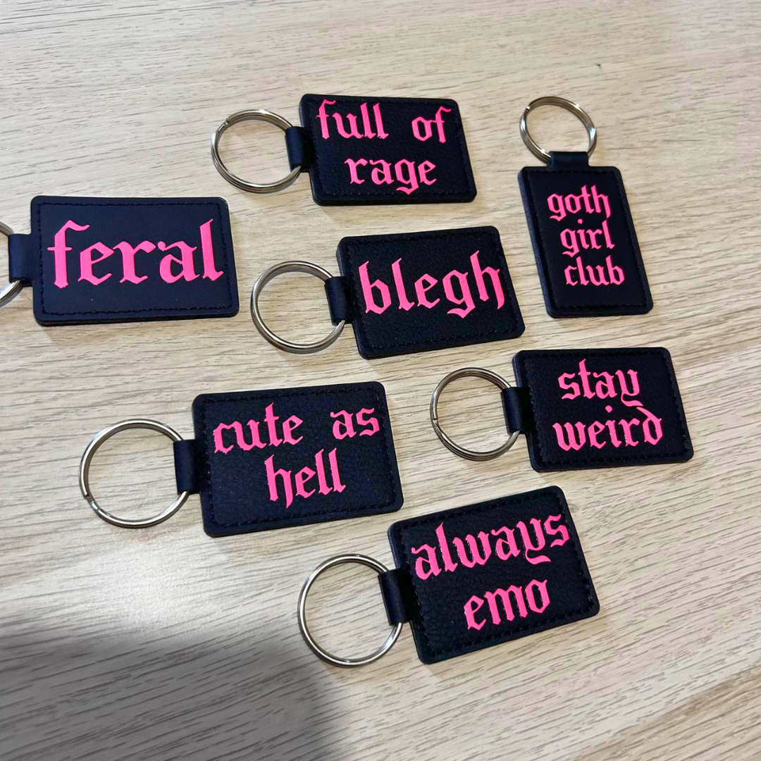 Seven black rectangular embroidered keychains with silver key rings, featuring pink Gothic-style text on a light wood background. The phrases include 'feral', 'full of rage', 'goth girl club', 'blegh', 'cute as hell', 'stay weird', and 'always emo'.