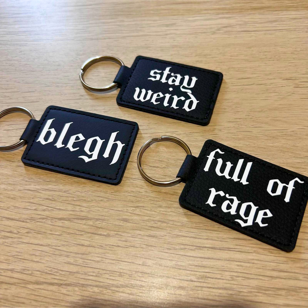 Three black rectangular keychains with white gothic text lying on a light wood surface. One keychain reads "stay weird," another reads "blegh," and the third reads "full of rage.