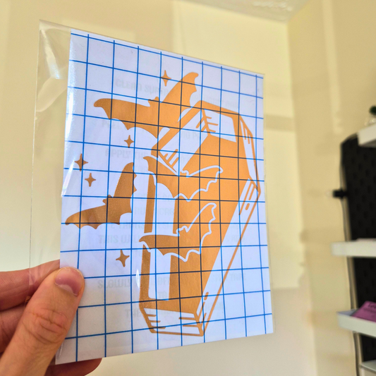 A hand holds a clear plastic sheet containing a printed orange coffin-and-bats graphic over a blue grid pattern, photographed indoors against a light wall.