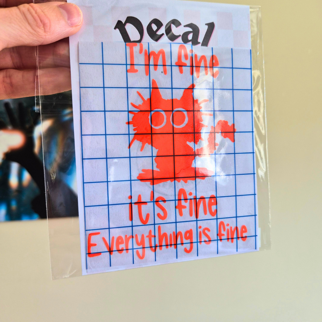 A hand holds a clear plastic bag containing a square decal sheet. The decal features the text 'Decal I'm fine it's fine Everything is fine' in orange-red lettering over a blue grid, with a red cartoon character wearing glasses in the center.
