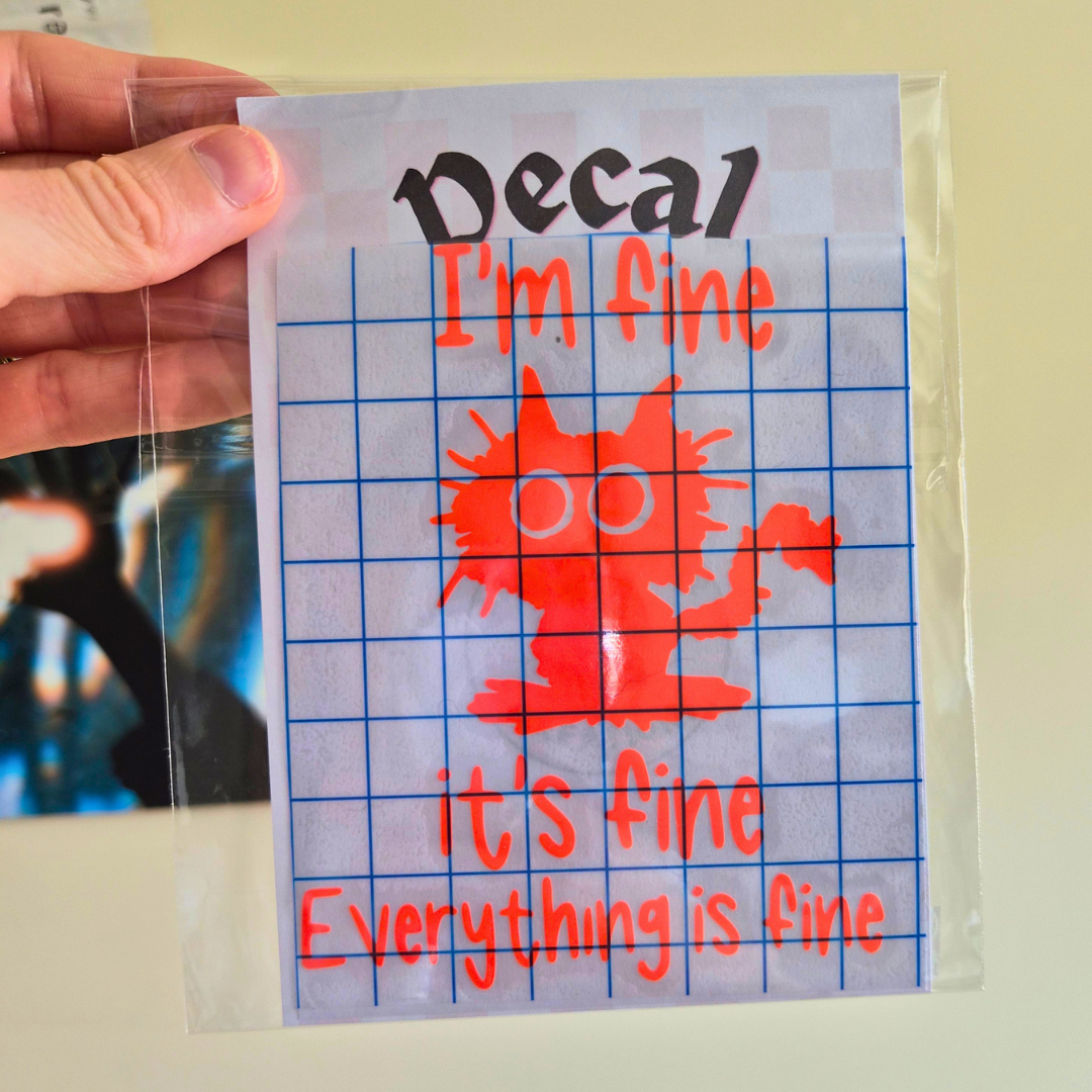 A hand holds a clear plastic bag containing a square decal sheet with the text 'Decal I'm fine it's fine everything is fine' and a red cartoon cat graphic on a white grid background.