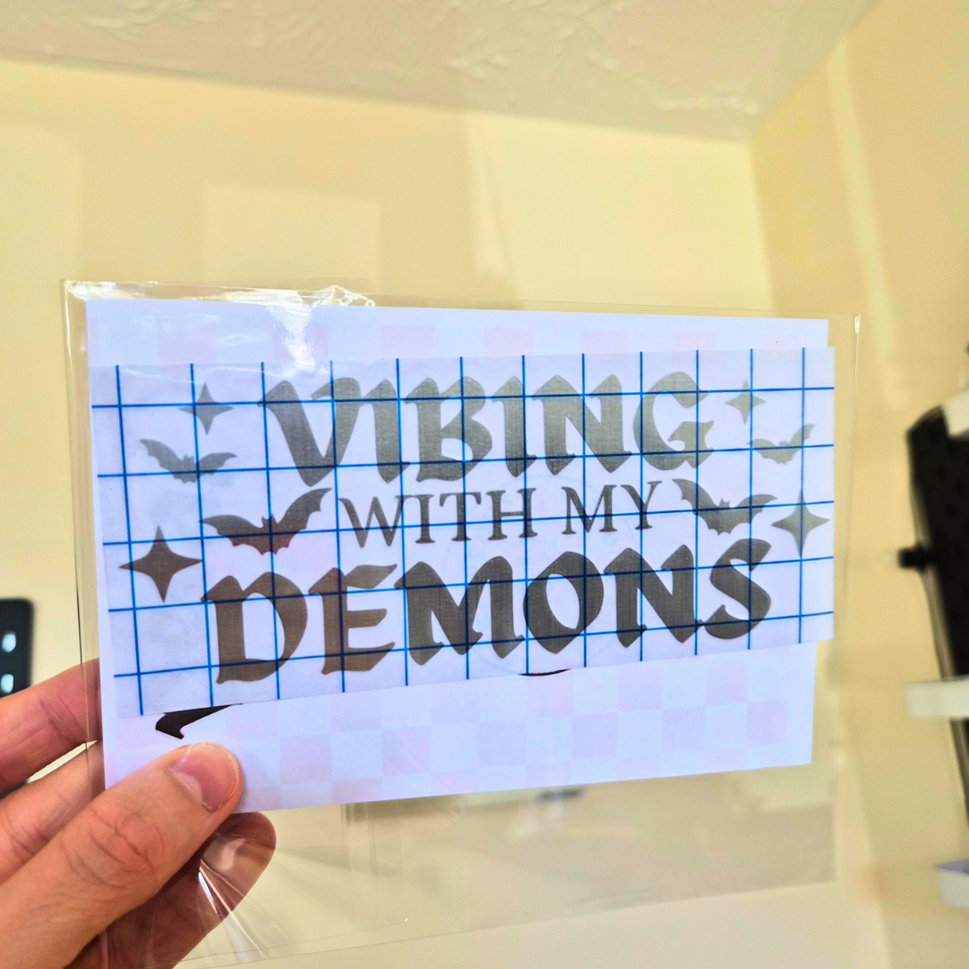 A hand holds a clear plastic-wrapped square card featuring the phrase “VIBING WITH MY DEMONS” in large dark gray lettering over a blue grid and bat illustrations. The background is a softly blurred indoor room with pale yellow walls.